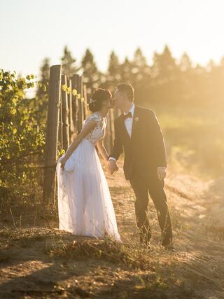 A couple in formal attire shares a tender kiss by a vineyard at sunset, surrounded by golden light and rustic fencing.