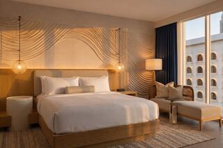 A stylish hotel room with a large bed, warm lighting, and modern decor, featuring a cozy seating area and large windows for sunlight.