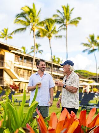 Two men enjoy drinks while chatting in a lush tropical setting with palm trees and vibrant flowers, surrounded by a relaxed atmosphere.