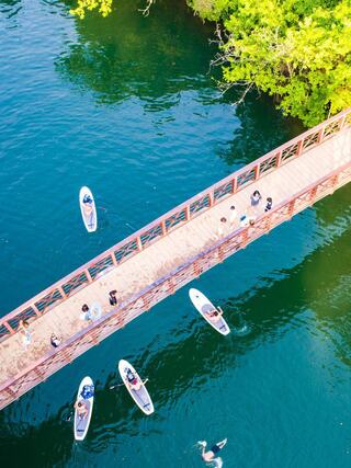 A scenic river scene features a wooden bridge and small boats navigating clear waters, surrounded by lush greenery and people enjoying the outdoors.