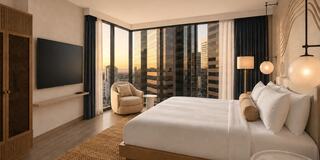 A modern hotel room features a large bed, cozy chair, and expansive windows offering a sunset view over city skyscrapers.