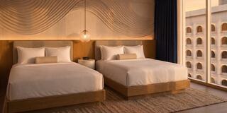 A stylish hotel room features two neatly made beds, warm wooden accents, soft lighting, and modern decor, creating a cozy atmosphere.