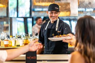 A smiling chef serves a plate of food at a vibrant bar, with patrons enjoying the lively atmosphere in the background.