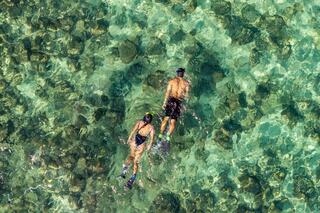 Two snorkelers explore clear turquoise waters, gliding over visible rocks beneath the surface in a serene, sunlit environment.