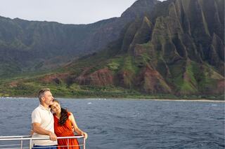 A couple embraces on a boat, surrounded by stunning green mountains and a calm sea, enjoying a serene moment together.