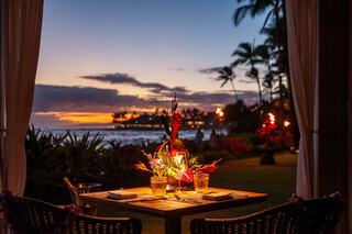 A cozy dining setup with two glasses and tropical flowers, framed by palm trees and a beautiful sunset over the ocean.