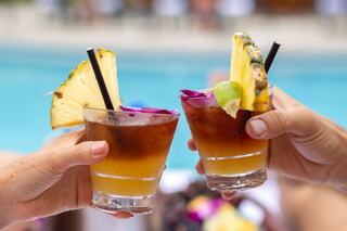 Two tropical cocktails clink together, garnished with pineapple and flowers, by a sparkling pool, evoking a relaxing, sunny vibe.