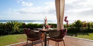 A cozy outdoor dining setup with two chairs overlooks a tranquil beach and vibrant tropical plants under a partly cloudy sky.