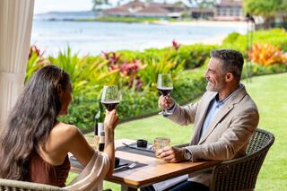 A couple enjoys a romantic meal by the ocean, with lush greenery in the background, creating a serene and intimate atmosphere.