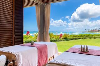 Two massage tables with soft linens sit in a serene spa overlooking a vibrant beach and lush greenery under a bright blue sky.