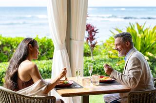 A couple enjoys a romantic meal by the ocean, surrounded by lush greenery and a serene atmosphere, sharing smiles and champagne.