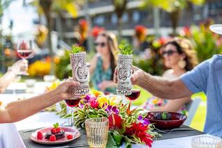 Tropical drinks clink at a vibrant outdoor gathering, surrounded by lush flowers and joyful guests enjoying a festive atmosphere.