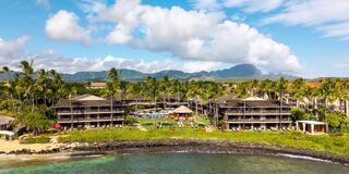 A tropical resort features lush palm trees, a pool area filled with sunbathers, and stunning mountain views under a bright blue sky.