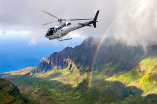 A helicopter soars over lush green mountains and a coastline, with a vibrant rainbow arching beneath cloudy skies.