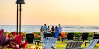 A group of people stands together, gazing at the ocean under a golden sunset, surrounded by vibrant flowers and empty lounge chairs.