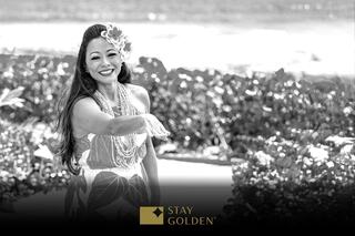 A smiling woman in a floral dress, adorned with a flower in her hair, gestures warmly amidst lush greenery, evoking a tropical vibe.