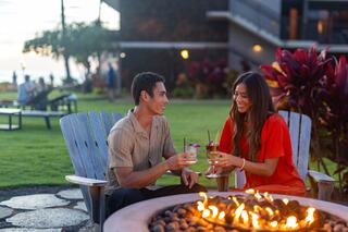 A couple enjoys cocktails by a fire pit, smiling at each other in a relaxed outdoor setting with lush greenery and a sunset in the background.
