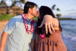 A couple kisses on the beach, showcasing an engagement ring, surrounded by tropical scenery and wearing leis.