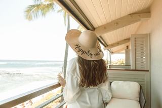A woman in a wide-brimmed hat stands on a balcony, overlooking a serene beach with palm trees swaying in the breeze.
