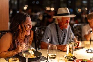 A woman laughs joyfully while enjoying dinner at a festive gathering, seated beside an older man in a hat and lei. Elegant ambiance surrounds them.