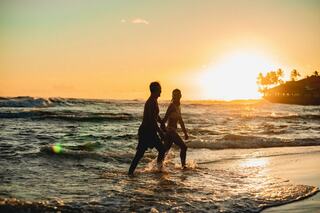 Silhouetted figures walk hand-in-hand along the shore, basking in the warm glow of a sunset over the ocean. Waves crash gently around them.