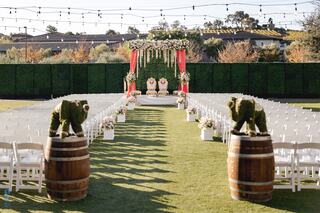 A beautifully decorated outdoor wedding aisle features white chairs, floral arrangements, and charming bear topiaries atop wooden barrels.