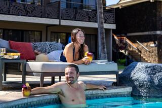 Two people relax by a pool, enjoying drinks. One lounges on a chair, while the other sits in the water, both smiling and enjoying the sunshine.