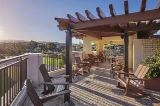 A sunlit terrace features wooden rocking chairs under a pergola, surrounded by greenery and scenic views, creating a relaxing atmosphere.