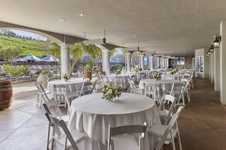 A spacious outdoor venue features round tables with white tablecloths, centerpieces, and views of lush greenery and an inviting pool area.