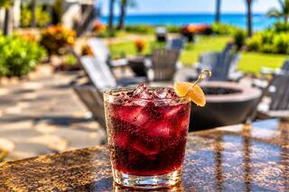 A refreshing cocktail with ice and a lemon twist sits on a table, overlooking a serene beach and lush garden.
