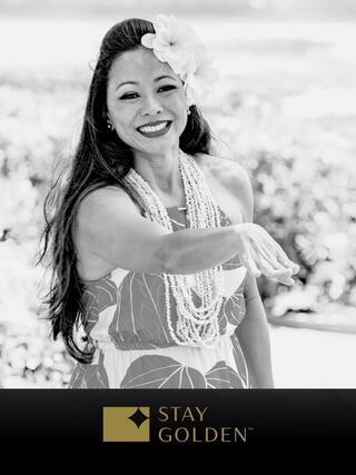 A smiling woman in a floral dress and necklaces extends her hand, exuding warmth and positivity, adorned with a flower in her hair.
