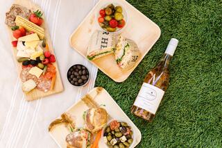 A picnic spread features assorted cheeses, fruits, sandwiches, pickles, and a bottle of wine, all set on lush green grass.