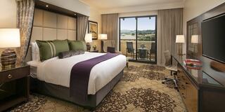 A cozy luxury hotel room features a large bed with plush pillows, a desk, lamps, and a balcony overlooking the scenery. Elegant decor enhances the ambiance.