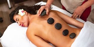A woman receives a soothing hot stone massage, with five smooth stones placed on her back and a flower in her hair, promoting relaxation.