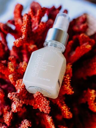 A bottle of Noni Skin Elixir is nestled among vibrant red coral, showcasing a sleek design and reflecting a natural beauty aesthetic.