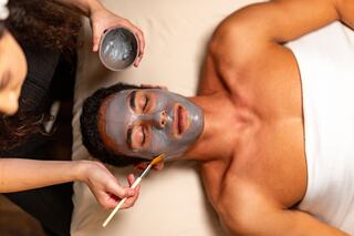 A man receives a facial treatment while lying on a table, eyes closed, as a person applies a mask with a brush. Relaxation in progress.