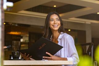 A woman with long hair smiles while looking at a menu in a cozy café setting, enjoying her time. A cup sits on the table nearby.