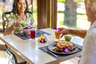 A cheerful couple enjoys breakfast together, featuring pancakes adorned with flowers and vibrant drinks, in a bright, inviting café.