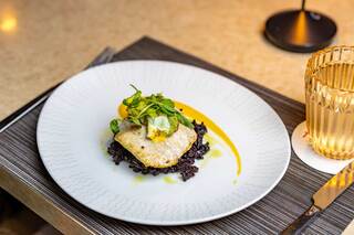 A beautifully plated dish featuring a piece of white fish atop black rice, garnished with greens and a vibrant sauce. A stylish glass is nearby.