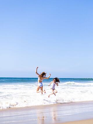 Two girls joyfully jump along a sandy beach, splashing in the waves under a clear blue sky. Bright and playful summer vibes.