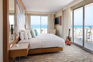 A modern hotel room featuring a large bed, stylish decor, and a view of the ocean through expansive windows, with a guitar in the corner.