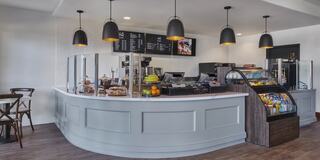A modern café features a curved counter, menu board, display of snacks, and fresh fruit, with stylish pendant lighting above.
