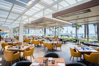 A vibrant restaurant with modern decor, featuring yellow chairs, wooden tables, and large windows offering a view of palm trees and the ocean.
