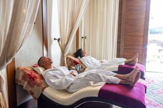 Two people relax on lounge chairs, wearing white robes, enjoying a serene spa environment with soft lighting and comfortable decor.