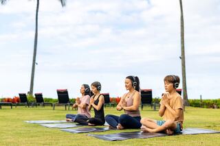 Four individuals practice yoga outdoors on mats, wearing headphones, surrounded by lush greenery and palm trees under a clear sky.