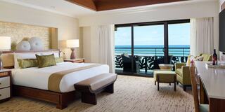 A luxurious hotel room featuring a king-sized bed, stylish seating, and a stunning ocean view through large glass doors.
