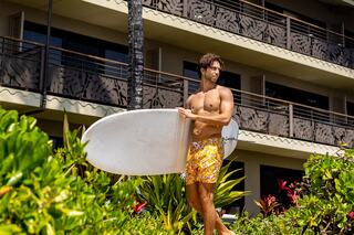 A shirtless man in vibrant shorts stands with a surfboard, surrounded by lush greenery and a tan building in the background.