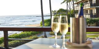 Two glasses of sparkling wine sit on a table, with an ocean view and palm trees in the background, creating a serene, tropical atmosphere.
