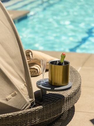 A sunbed beside a shimmering pool, featuring a chic gold champagne bucket, a glass, and neatly rolled towels, exuding relaxation.