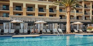 A serene poolside scene features loungers under umbrellas, palm trees, and a stylish hotel facade, perfect for relaxation.